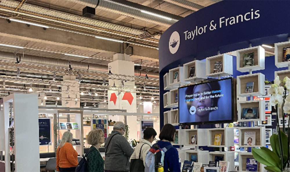 Visitors explore the Taylor & Francis exhibition booth at a book fair, featuring a curved blue display wall with books, a digital screen, and branded signage under an industrial ceiling with exposed ducts.