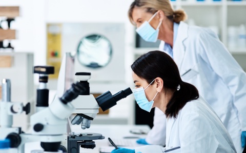 A photograp of two women wearing lab coats and masks. One is looking into a microscope.