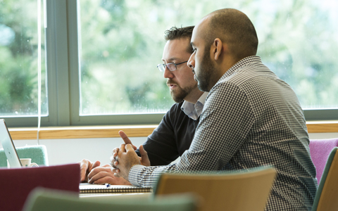 Two men sitting at a table looking at a laptop screen. One man is typing.